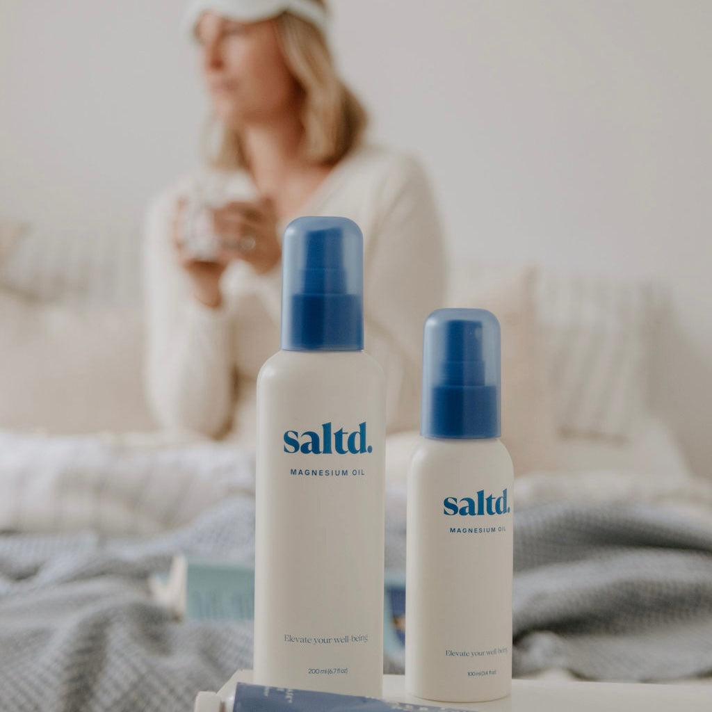 Two bottles of saltd magnesium oil on a surface with a blurred background of a person holding a mug.
