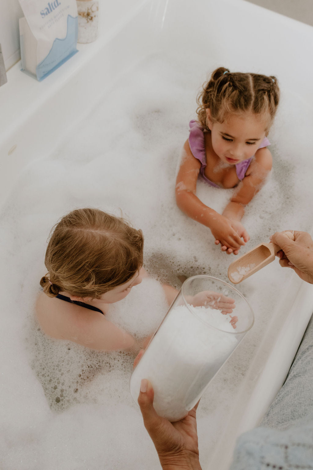 Two children in a bubble bath with a person pouring a magnesium flakes from a glass.