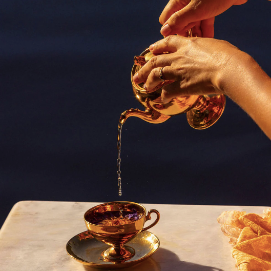 Gold teapot pouring blue lotus tea into a gold teacup