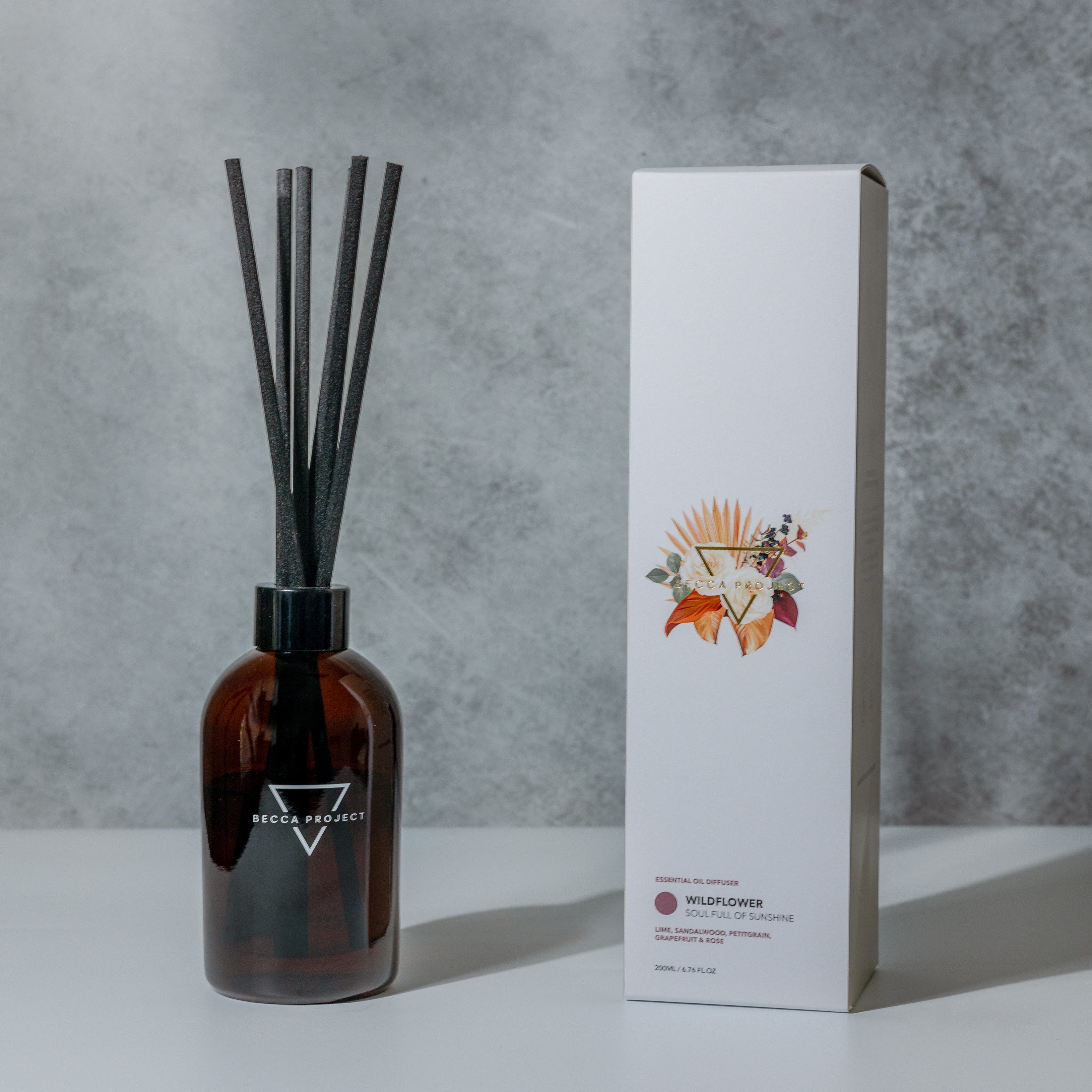 A Becca Project reed diffuser bottle with black reeds and a white box with floral design next to it, labeled 'Wildflower'.