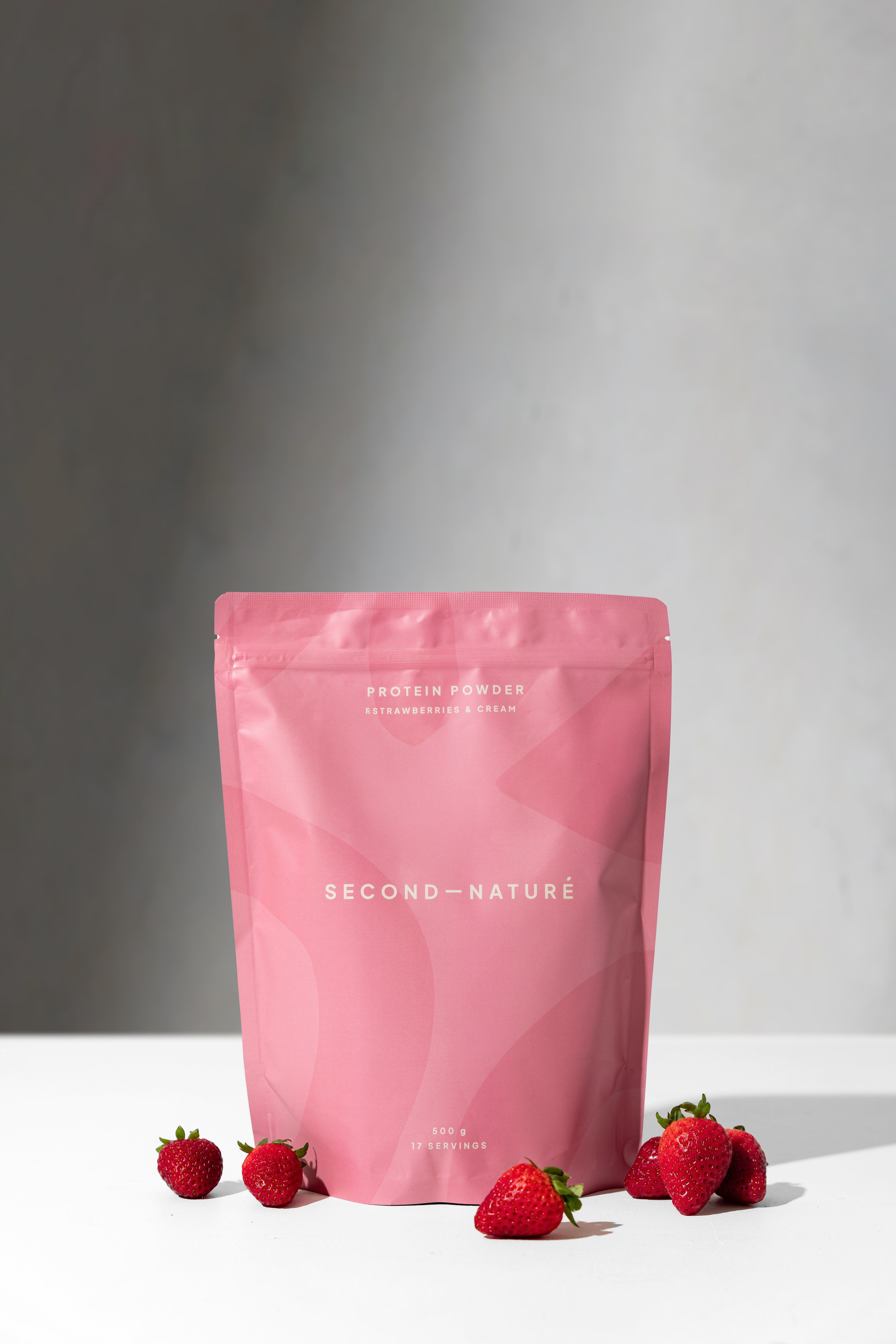 Second Nature Pink protein powder bag with strawberries on a white surface and grey background