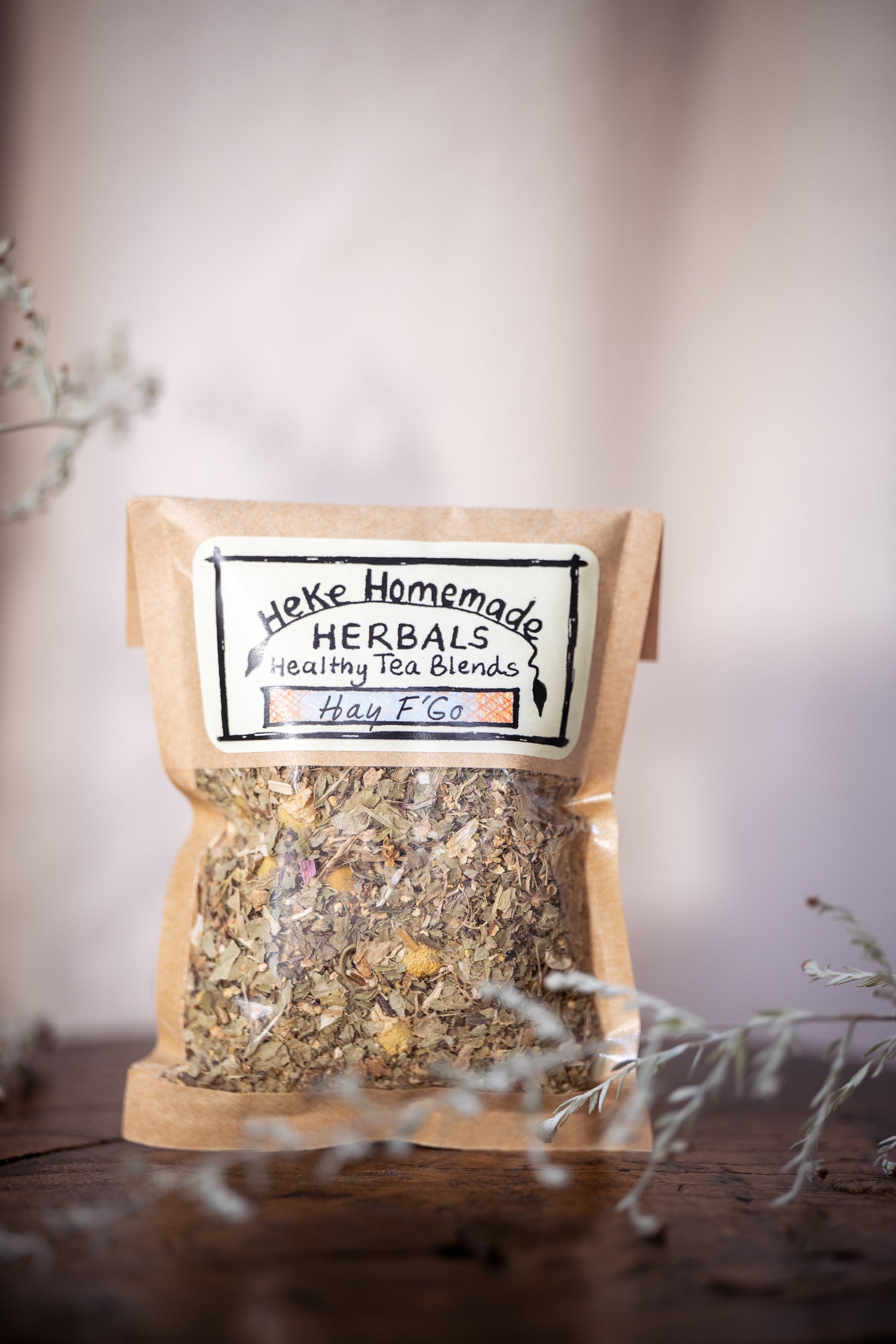 Packaged tea blend labeled 'Hay F'Go' from Heke Herbals, visible through a glass with a blurred background.