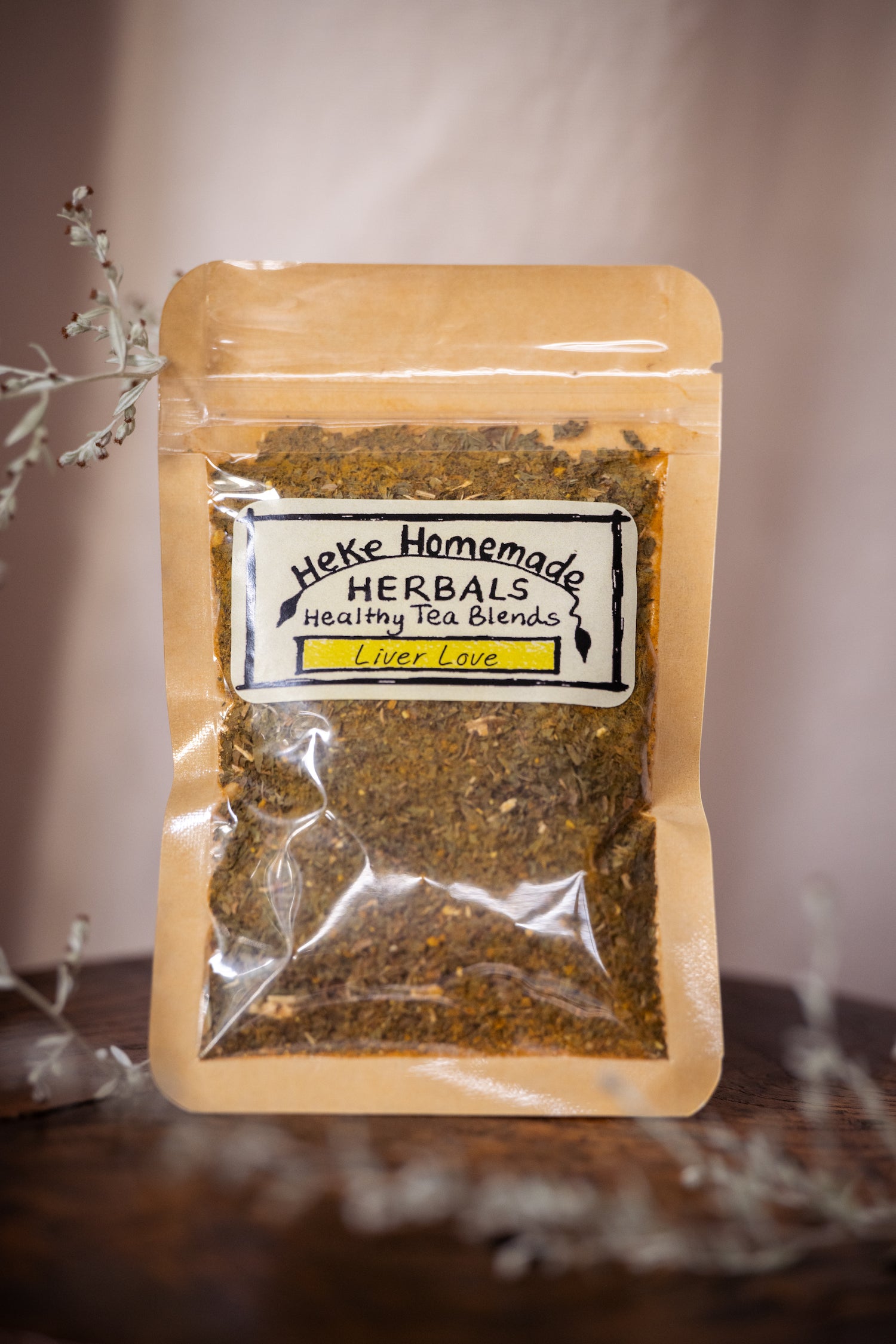 Packaged tea blend labeled 'Heke Herbals - liver love' with visible herbs through the clear packaging, on a dark background with decorative elements.