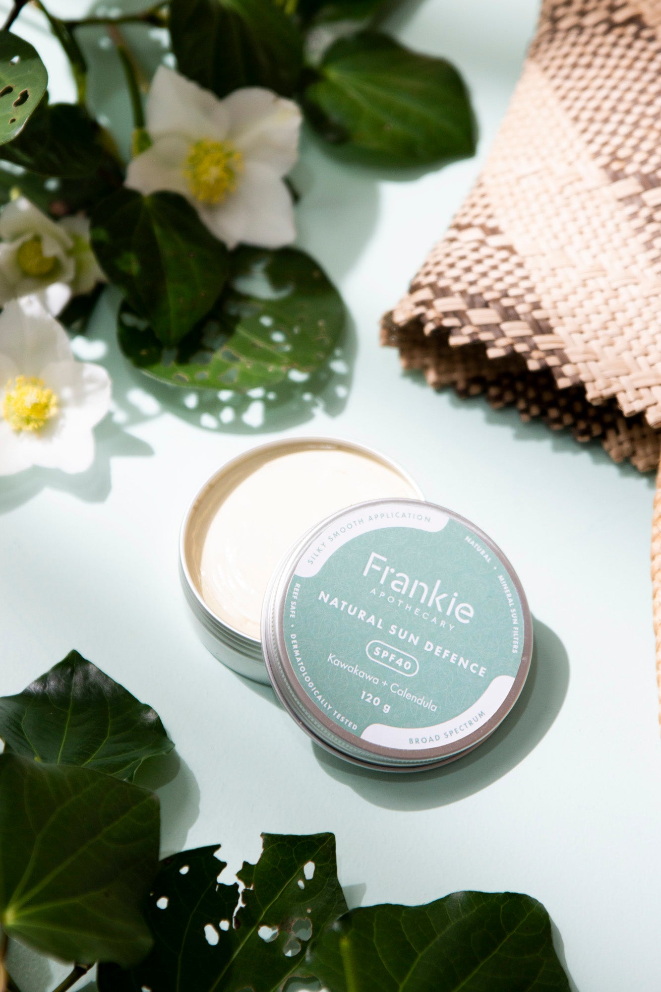 A compact container of Frankie Natural Sun Defence SPF40 sunscreen, with the product name and SPF level visible, displayed on a surface with green leaves and a floral background.