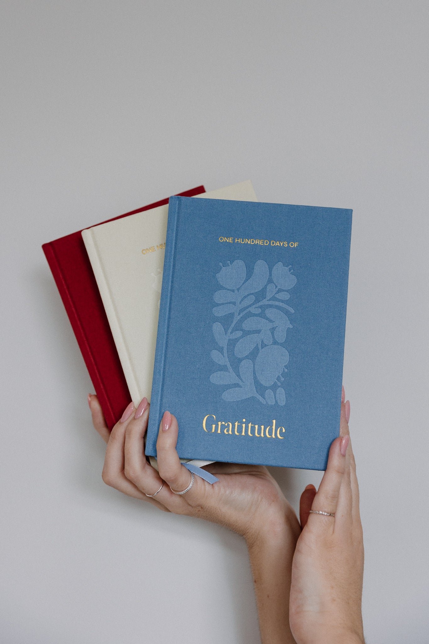 hands holding up three gratitude journals in wine, buttermilk and sky blue