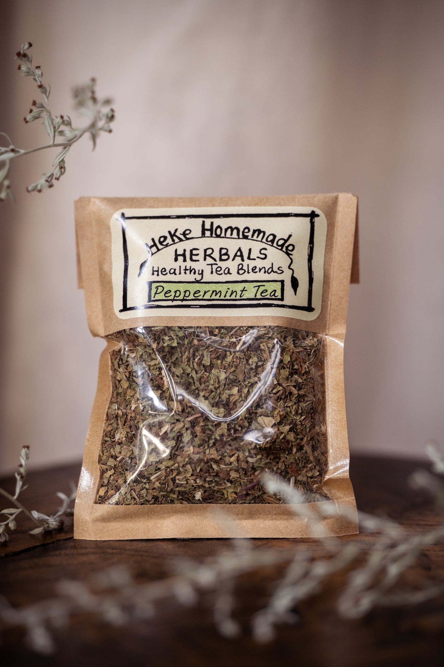 Packaged peppermint tea leaves in a clear bag with a beige label reading 'Heke Homemade Herbals Peppermint Tea', set on a dark background with some decorative elements.