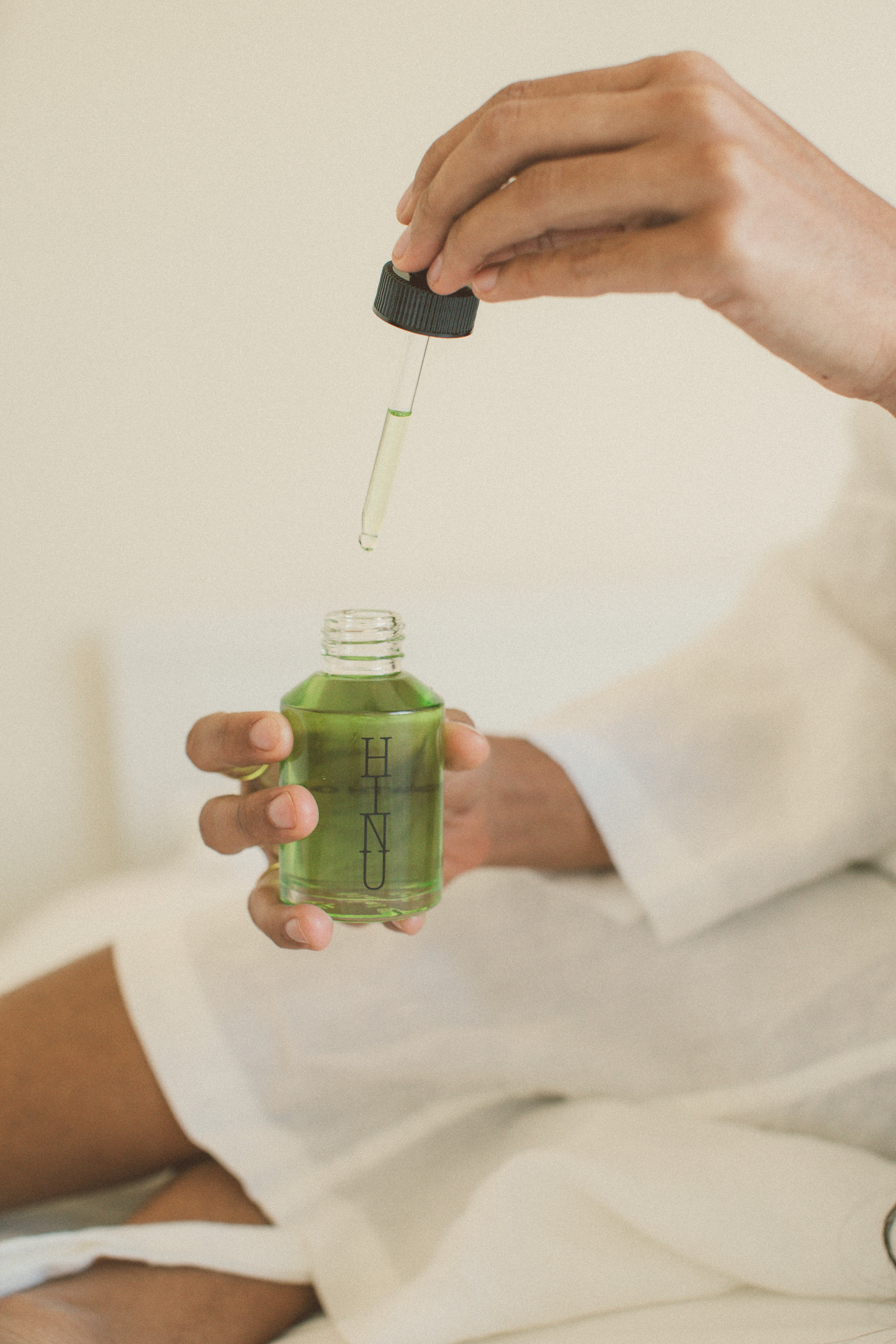 Person holding a dropper bottle of HINU hair growth oil, wearing a white robe.